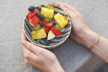 Hands holding a patterned bowl filled with fresh fruit skewers, including strawberries, pineapple, grapes, and blueberries, placed on a textured gray tablecloth. A simple, healthy snack option.