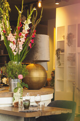 A stylish café interior with a round table featuring a tall gladiolus flower arrangement in a metal bucket, a large gold lamp, and a small wooden dining table set with wine glasses in the foreground.