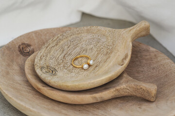 A gold ring with two pearls rests on stacked light wood dishes with a natural grain and "Handmade" branding. The image features warm, earthy tones, highlighting the minimalist and artisanal design.