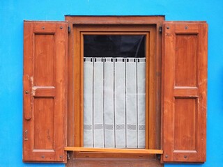 One isolated rustic window set in a blue facade, with open wooden shutters and a white lace curtain inside