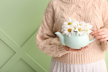Beautiful young woman with teapot of chamomile tea and flowers near green wall
