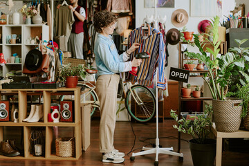 Woman organizing striped shirt display in chic boutique filled with various accessories, plants, and decor items, while another individual browses in the background