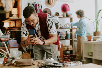 Two men engaging in activities within a retail environment. Focused individual is capturing a moment on his smartphone while the other arranges merchandise in background