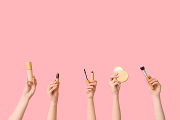 Female hands with makeup foundation, lipstick, mascara, highlighter and brush on pink background