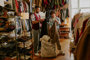 Two men browsing vintage clothing store's collection, holding different garments while discussing...