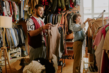 Man and woman browsing clothes and selecting items in vintage clothing store with racks full of colorful outfits surrounding them and lamp lighting the space