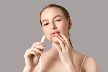 Beautiful young woman applying lip balm on grey background