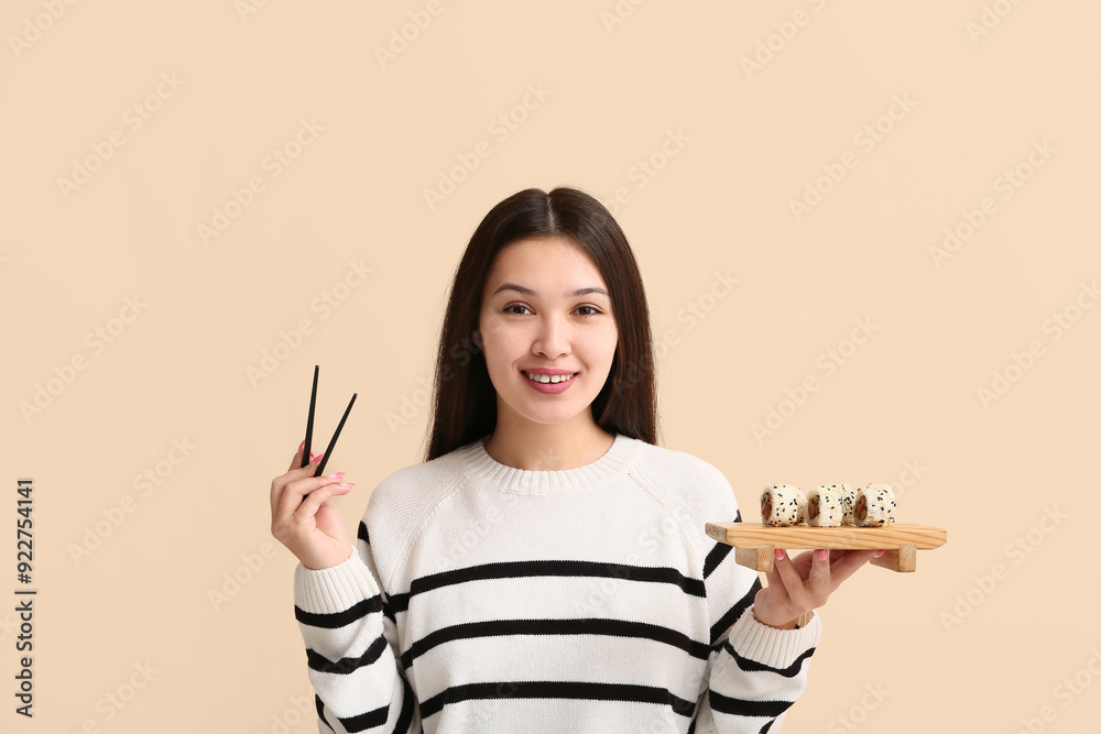 Sticker Beautiful young Asian woman holding chopsticks and wooden board with delicious sushi rolls on beige background