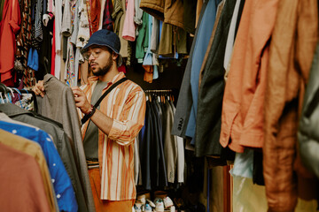 Young man with glasses and hat selecting clothes in a vibrant store with many options on both sides of aisle. He is carefully examining a grey jacket while carrying a crossbody bag