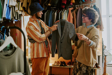 Two friends examining vintage jacket in cozy, well-stocked clothing store. Shelves filled with various clothes seen in background, natural light from large window illuminating scene