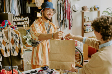 Smiling man wearing glasses and a hat handing shopping bag in a thrift shop with various clothes...