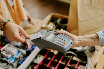Hand holding payment card while completing transaction through point-of-sale device in retail setting involving jewelry display. Close-up showing customer paying using electronic payment method