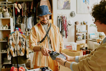 Smiling man wearing hat and glasses making payment at clothing store counter with cashier accepting...