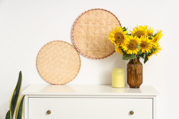 Vase of beautiful sunflowers bouquet with candle on commode near white wall at home, closeup