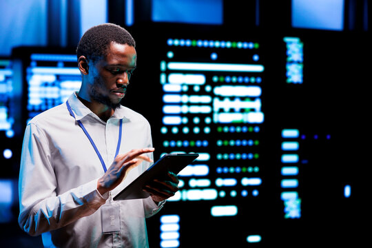 Computer scientist running high tech data center components diagnostic test to determine and patch software issues. African american employee using tablet to mend critical systems checking for defects