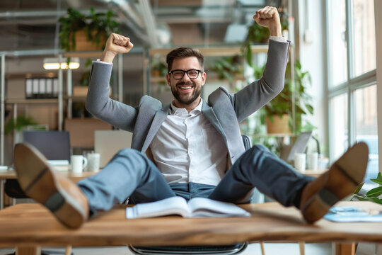 Euphoric businessman celebrating work success with fists up and feet on the table in an office with wooden furniture