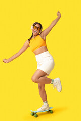 Emotional African-American woman with skateboard listening to music on yellow background