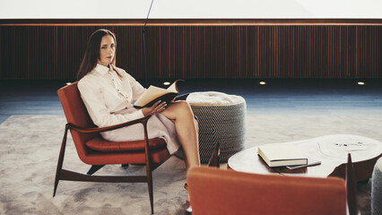 A hispanic woman is seated on an orange chair in a minimalist interior, holding an open book. The room is spacious with soft lighting, a modern design, and neutral tones - clean lines and simple decor