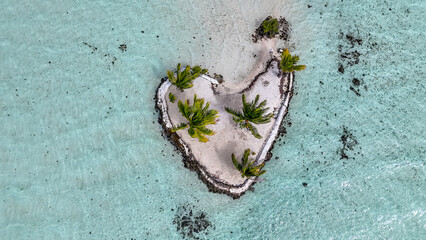 Aerial drone view of a heart shaped tropical island near Taha'a in French Polynesia. The white sand covered motu has several palm trees and is surrounded by azure and turquoise clear blue ocean water.