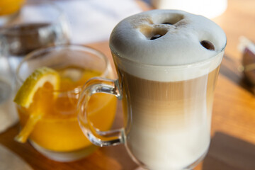 Hot latte in a steaming mug topped with foam and coffee beans on a breakfast table with fresh squeezed orange juice. 