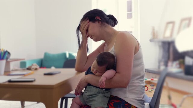 Mother sits at the table, head in hand, cradling a sleeping baby, embodying the fatigue and stress of motherhood, in a moment of quiet overwhelm in a home environment