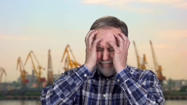Portrait of extremely upset mature man holding his head with both hands. Worried caucasian old male. Cranes and evening sky in the background.
