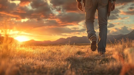 A person strolls through a grassy field under a vibrant sunset, with mountains silhouetted in the background.