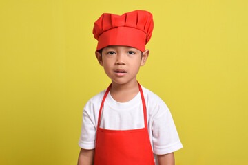 Cute little chef isolated on yellow background. Asian boy wearing red apron and hat.