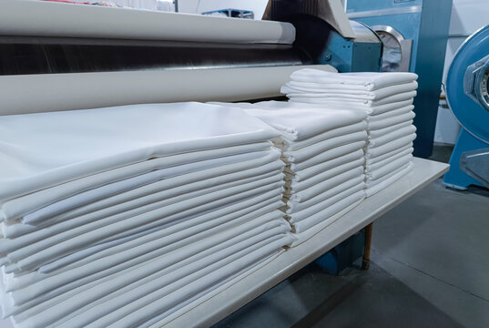 Folded white clothes, sheets, tablecloths, in an industrial laundry with machinery behind.