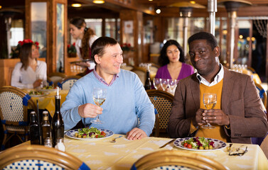 Two cheerful men having friendly conversation at dinner in cozy restaurant, sitting at served table and drinking wine..