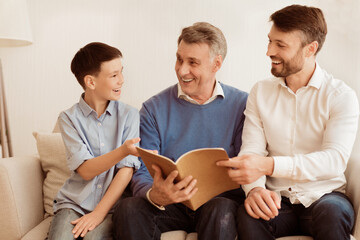 Obraz premium Male Family Of Three Reading Book Sitting On Sofa Indoor. Son, Father And Grandfather Spending Time Together. Selective Focus