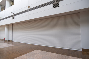 A large, wide, closed, and locked roller door of a shop in a modern indoor building. Commercial retail area with a blank white shutter gate. Copy space for design.