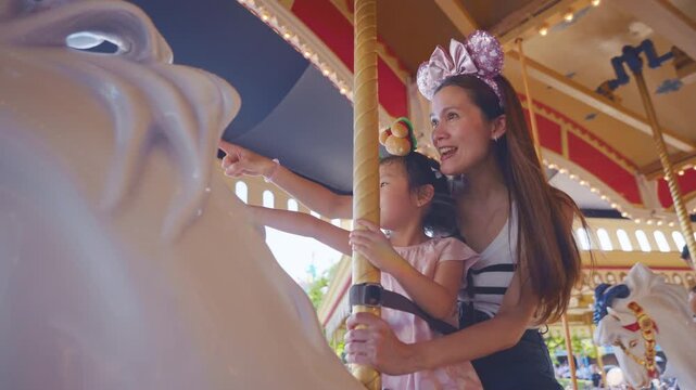 Asian mother and adorable kid daughter riding carousel at theme park. 