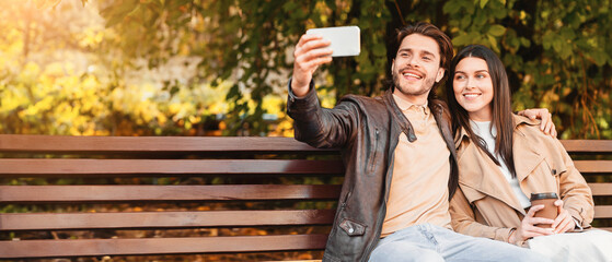 Happy couple in love taking selfie on smartphone, sitting on bench in park