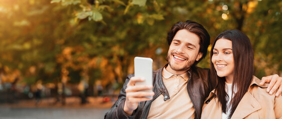 Young happy lovers enjoying time together, taking selfie while walking by golden forest, using mobile phone, close up