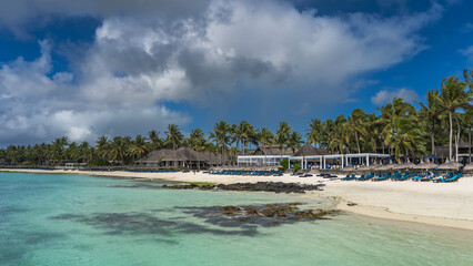 On the sandy beach by the turquoise ocean there are rows of sun loungers under straw umbrellas. The thatched roofs of the hotel buildings are surrounded by tall palm trees. Blue sky, clouds. Mauritius
