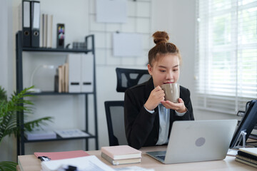 Naklejka premium A confident businesswoman in a black suit is sitting at her desk, smiling as she works on her laptop, showcasing a modern and professional workspace