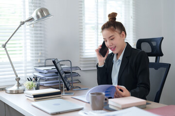 A businesswoman in a black suit is talking on the phone while working at her desk with a laptop, embodying multitasking and efficiency in a professional office setting