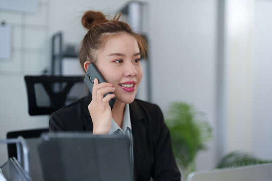 A businesswoman in a black suit is talking on the phone while working at her desk with a laptop, embodying multitasking and efficiency in a professional office setting