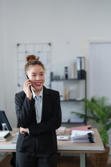 A businesswoman in a black suit is standing near her desk, talking on the phone, exuding confidence and professionalism in a modern office environment