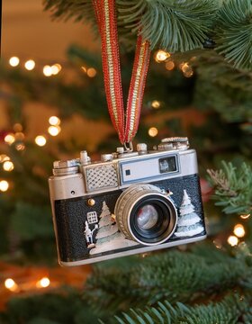 A classic camera ornament featuring white Christmas trees and a red ribbon, hanging on a Christmas tree. Ideal for use in nostalgic holiday advertisements or vintage-themed decorations.