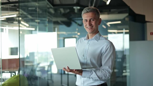 Confident businessman holding laptop and smiling at camera in modern office. Working on Design, Data Analysis, Plan Strategy. Adult happy man leader boss professional manager model posing at workplace
