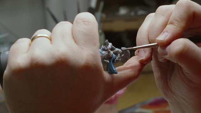 Artist Holding Miniature Fantasy Figure Model Tightly Whilst Painting Delicately with Paint Brush in Art Studio 4K