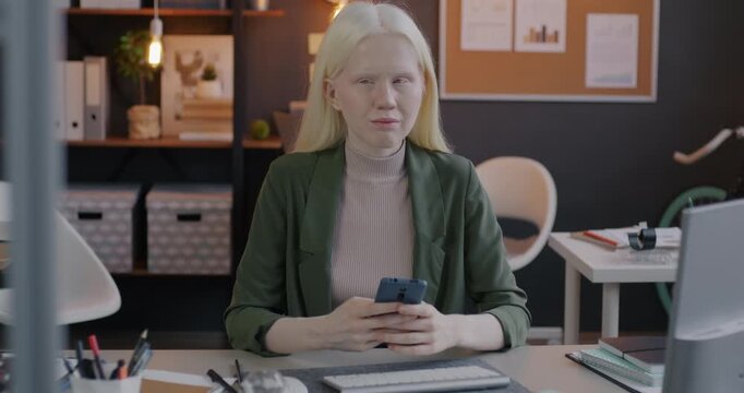 Slow motion portrait of young albino businesswoman using smartphone then looking at camera at desk in office. Corporate lifestyle and inclusive workplace concept.