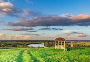 A small wooden gazebo sits in a grassy field with a river in the background