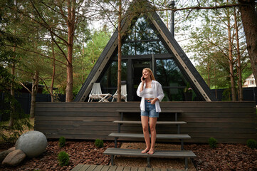 Young beautiful European woman in white shirt and jean shorts talking on the phone standing on the steps of A-frame house on a sunny summer afternoon.