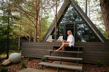 Enjoying free time. A happy European woman reading an interesting book sitting on the steps of a country a-frame house on a sunny day, free space