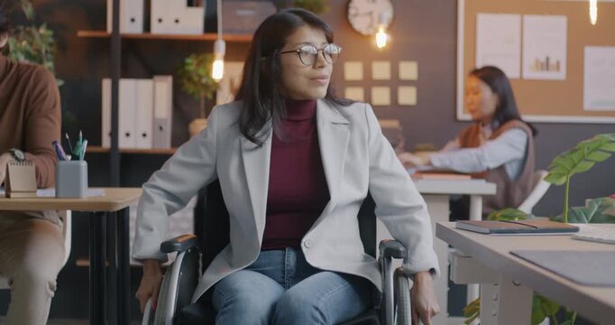 Slow motion of young Middle Eastern woman in wheelchair moving in inclusive workspace while colleagues working at desks in office. Job and disabled employee concept.
