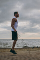 Young strong sporty athletic toned fit sportsman man he wears sports clothes jumping on skipping rope warm up training at sunrise sun dawn over sea beach outdoor on pier seaside in summer day morning.