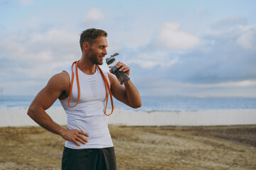 Young strong sporty athletic toned fit sportsman man he wear sports clothes elastic rubber band, drink water warm up train at sunrise sun dawn over sea beach outdoor on seaside in summer day morning.